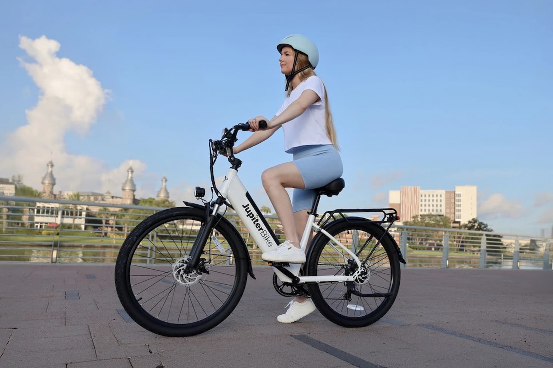 JupiterBikes Cruise Electric Bike – A woman wearing a white helmet, white t-shirt, and light blue shorts riding a white and black electric bicycle with black tires on an outdoor paved path under a blue sky.