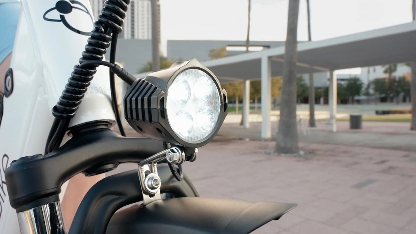 JupiterBikes Cruise Electric Bike – A close-up shot of a black LED motorcycle front light mounted on a handlebar, with a blurred outdoor urban background featuring a covered walkway and palm trees.