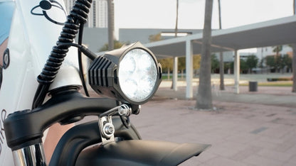 JupiterBikes Cruise Electric Bike – A close-up shot of a black LED motorcycle front light mounted on a handlebar, with a blurred outdoor urban background featuring a covered walkway and palm trees.
